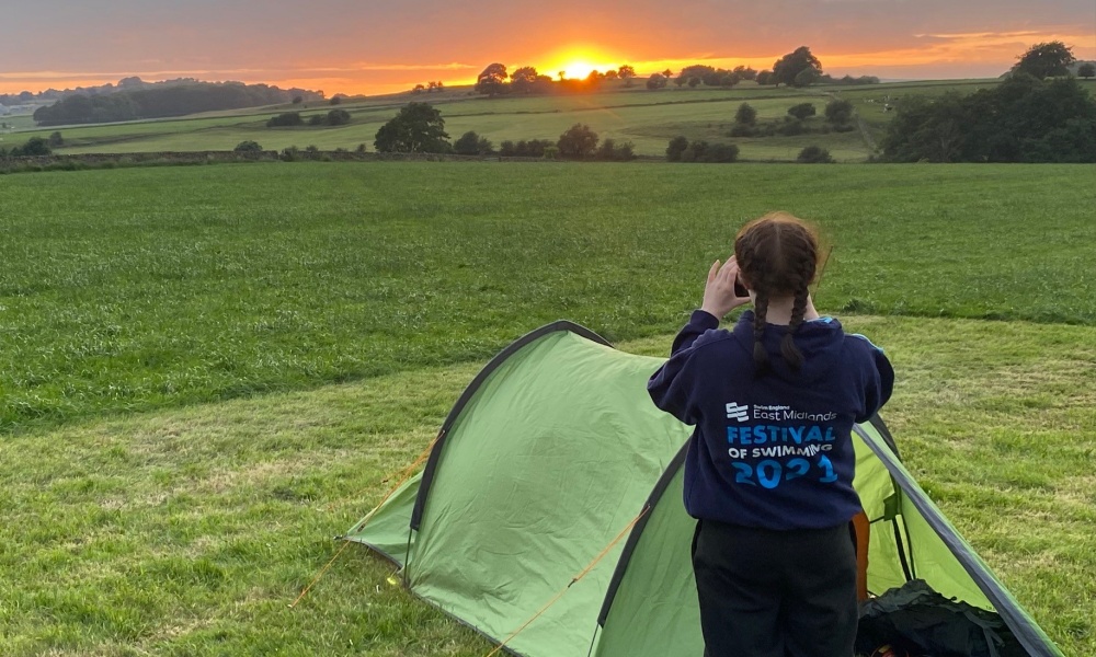 A student standing next to her tent taking a photo of the sunset